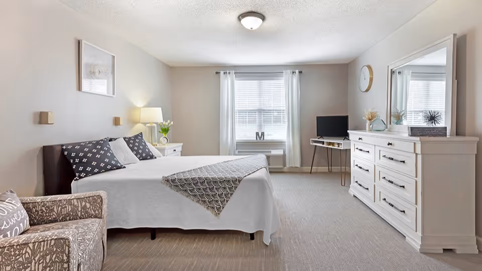 A bright and spacious bedroom with a large bed covered in white and patterned bedding, a patterned armchair with a pillow, a white nightstand with a lamp and flowers, a window with white curtains, a small desk with a letter M decoration, a TV on a stand, and a white dresser with a mirror and decorative items.