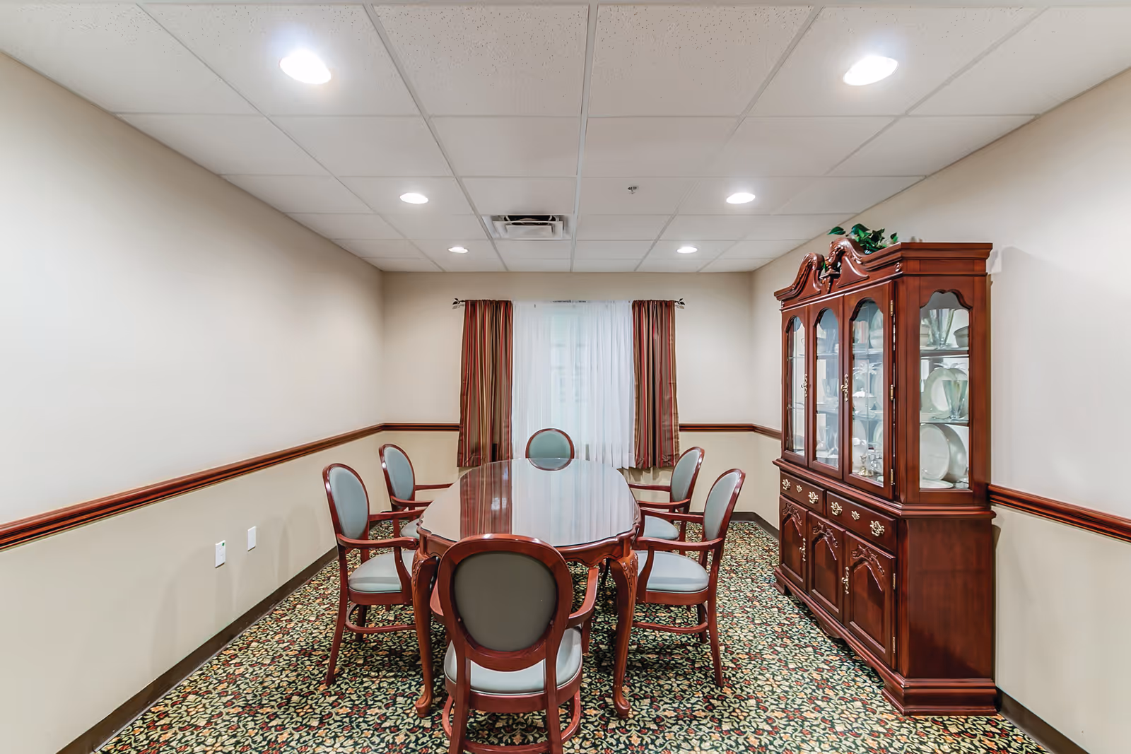 A small dining room with a polished wooden oval table surrounded by six chairs with green cushions. There is a large wooden china cabinet filled with plates and glassware against the right wall. The room has beige walls, a patterned carpet, a window with sheer white curtains and red drapes, and a drop ceiling with recessed lighting.