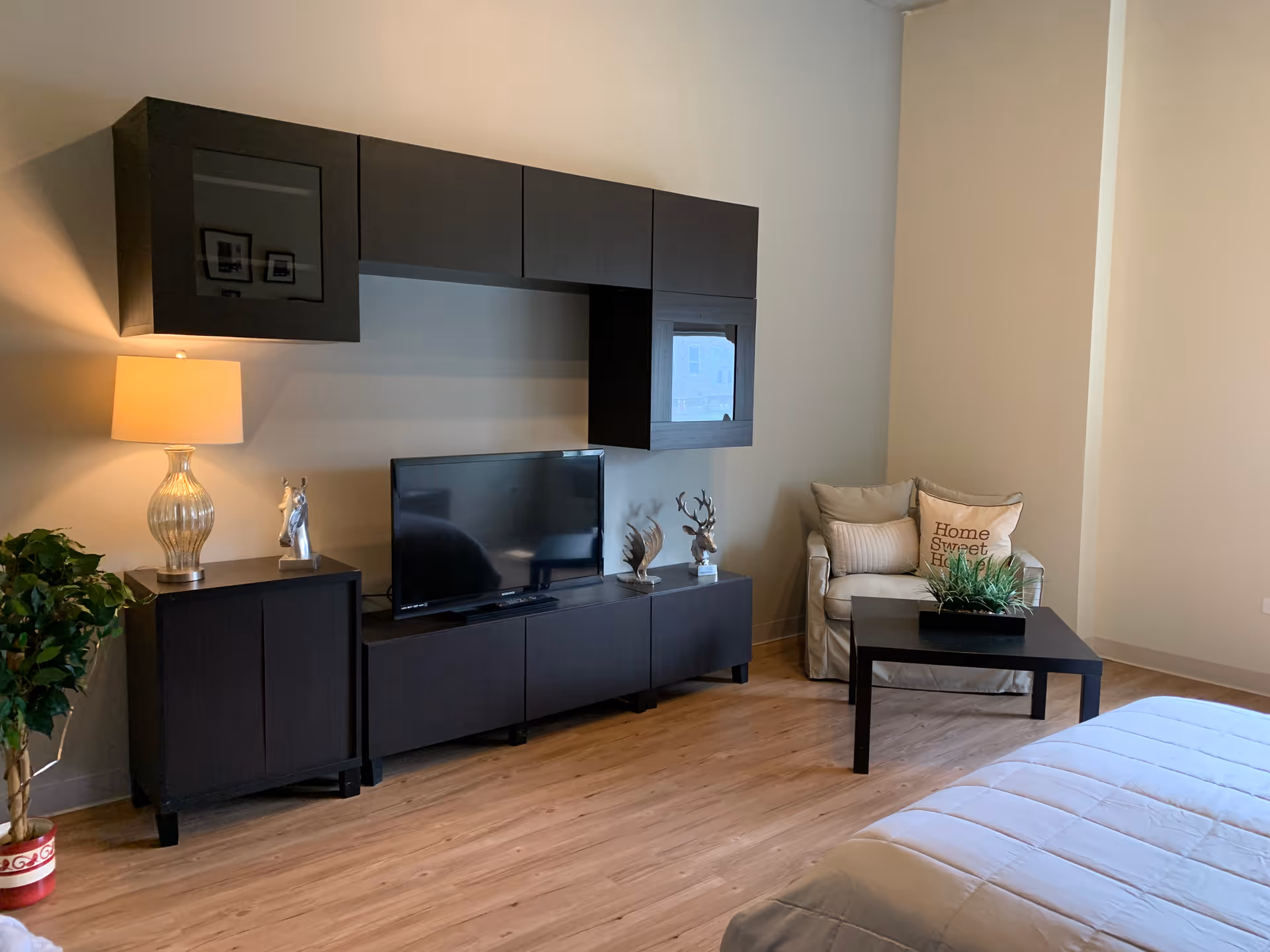 A cozy living room area with a dark wood entertainment center holding a flat-screen TV, decorative items, and a lamp. To the right, there is a cushioned armchair with a pillow that reads 'Home Sweet Home' and a black coffee table with a green plant on it. The floor is wooden, and part of a bed with a white comforter is visible in the foreground.