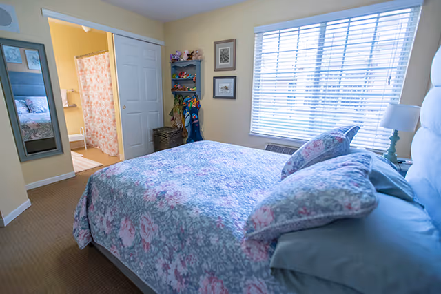 A cozy bedroom with a bed covered in a floral patterned comforter and pillows. There is a window with blinds letting in natural light, a bedside table with a lamp, and a wall-mounted mirror. An open door reveals a bathroom with a floral shower curtain and a glimpse of a toilet.