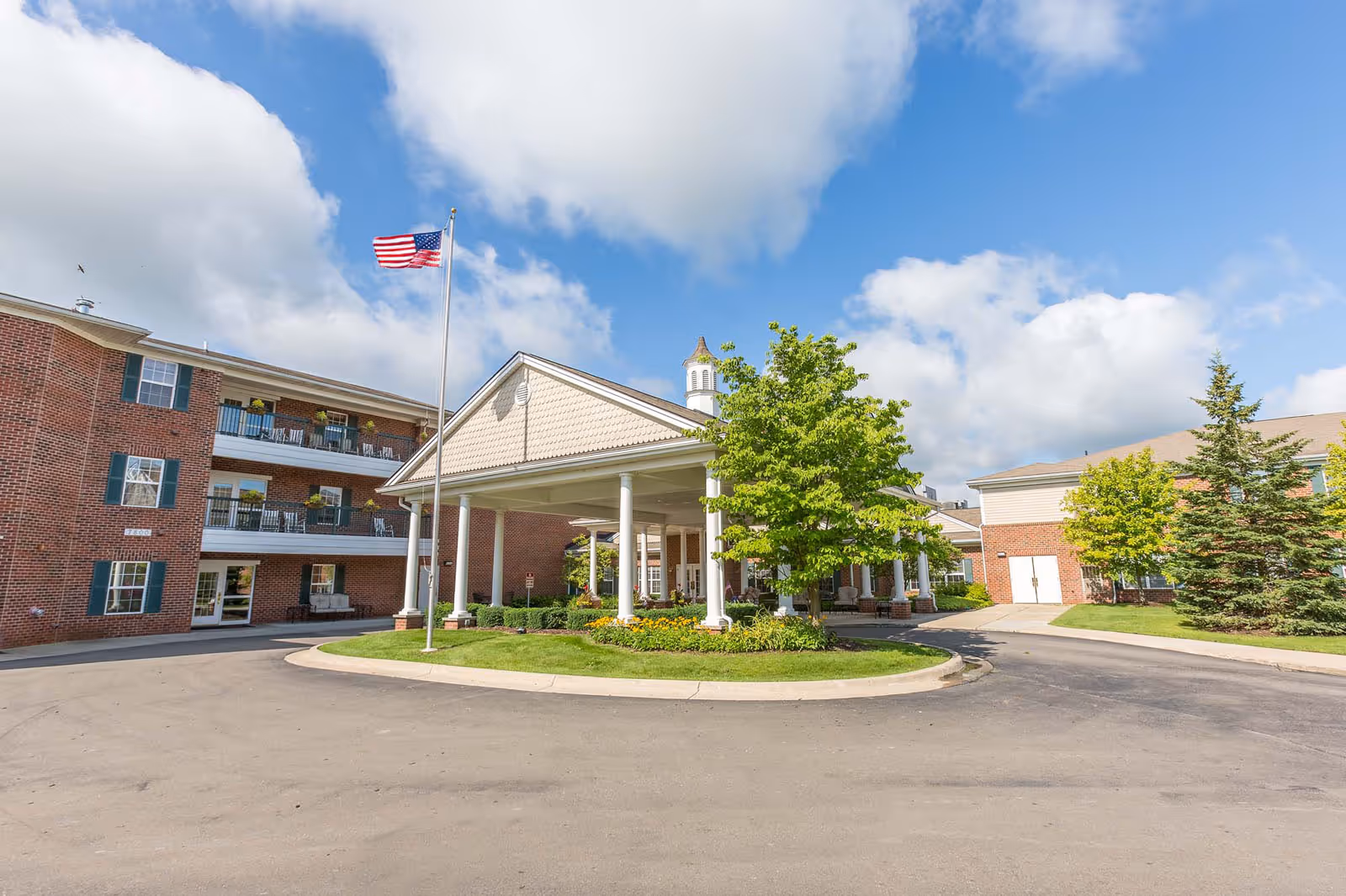 Front entrance of a brick senior living building with a covered porte-cochere, American flag, and landscaped circular driveway.