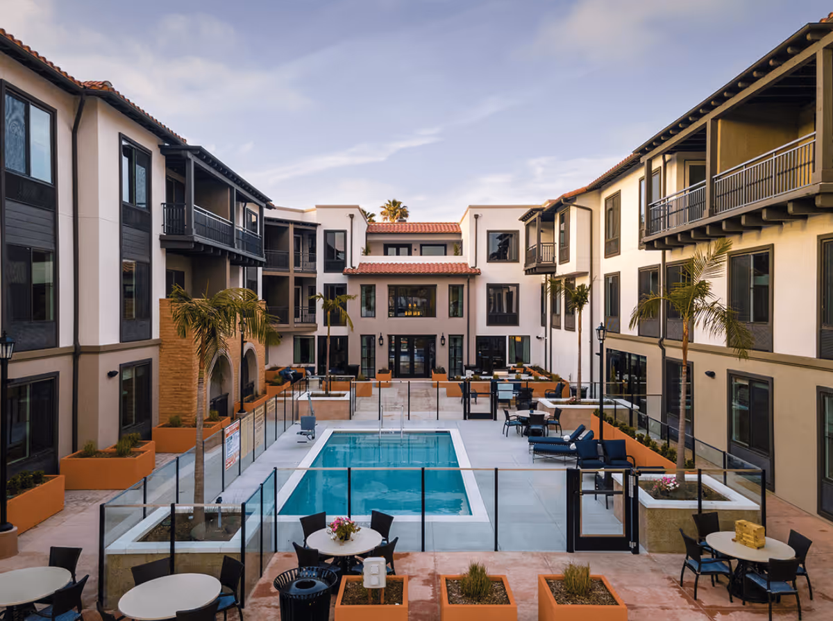 Outdoor courtyard area of a senior living facility featuring a rectangular swimming pool surrounded by glass fencing. The courtyard is flanked by three-story buildings with balconies, palm trees, and multiple seating areas with tables and chairs. The sky is partly cloudy.