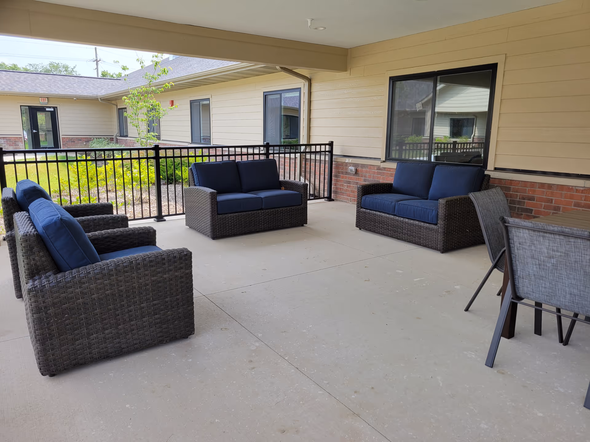Covered outdoor patio area with wicker furniture including two loveseats and two armchairs with blue cushions, a table with four chairs, and a black metal railing overlooking a garden area. The building exterior has beige siding and brick accents with several windows.