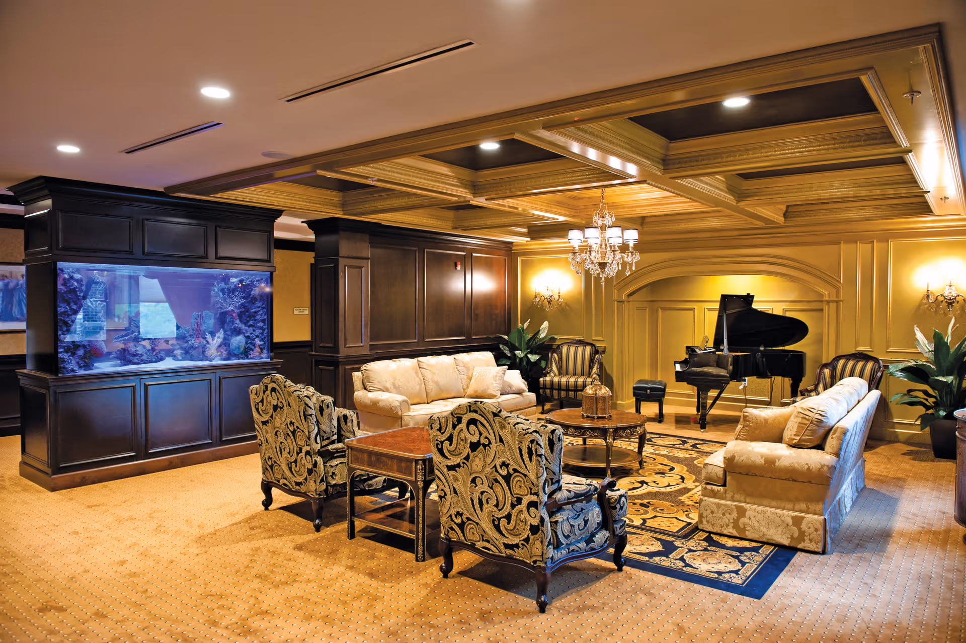 A luxurious living room with beige patterned sofas and armchairs arranged around a wooden coffee table on a decorative rug. There is a large built-in aquarium on the left side and a grand piano with a piano bench in the background. The room features a coffered ceiling with recessed lighting and a chandelier, along with wall sconces and potted plants.