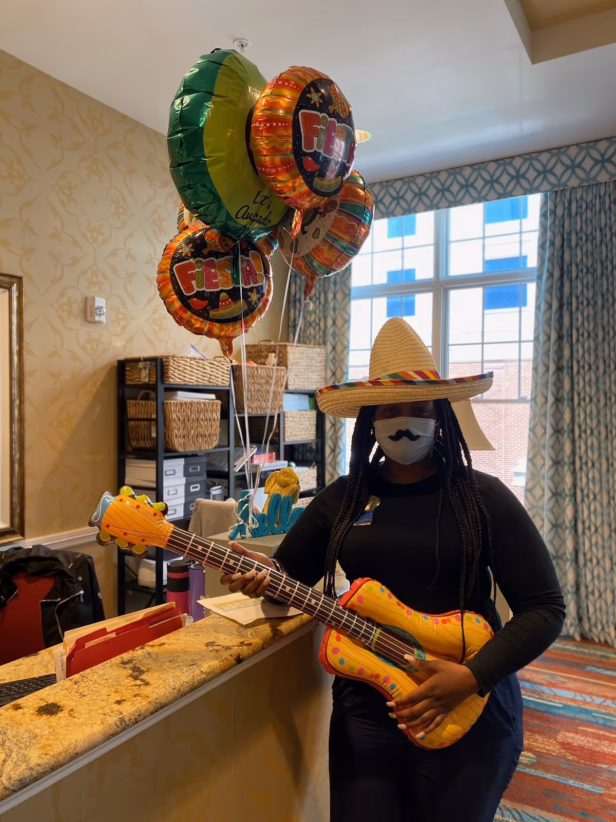 A person wearing a sombrero hat and a face mask with a mustache design is standing behind a reception desk holding an inflatable guitar. Behind them are colorful Fiesta-themed balloons and shelves with baskets. The room has patterned curtains and a large window letting in natural light.