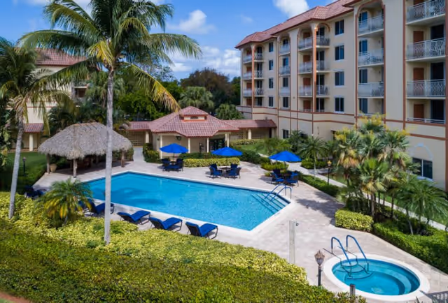 Landscaped courtyard with a swimming pool, hot tub, lounge chairs and umbrellas next to a multi-story residential building.