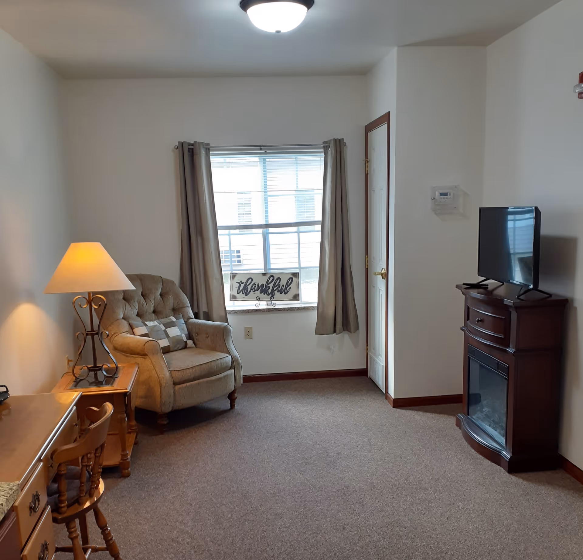 A cozy living room area with a beige armchair next to a wooden side table with a lamp. A window with beige curtains and a decorative sign that reads 'thankful' is centered on the wall. To the right, there is a dark wooden TV stand with a flat-screen TV on top and an electric fireplace below. The room has beige carpet and white walls with wooden trim.