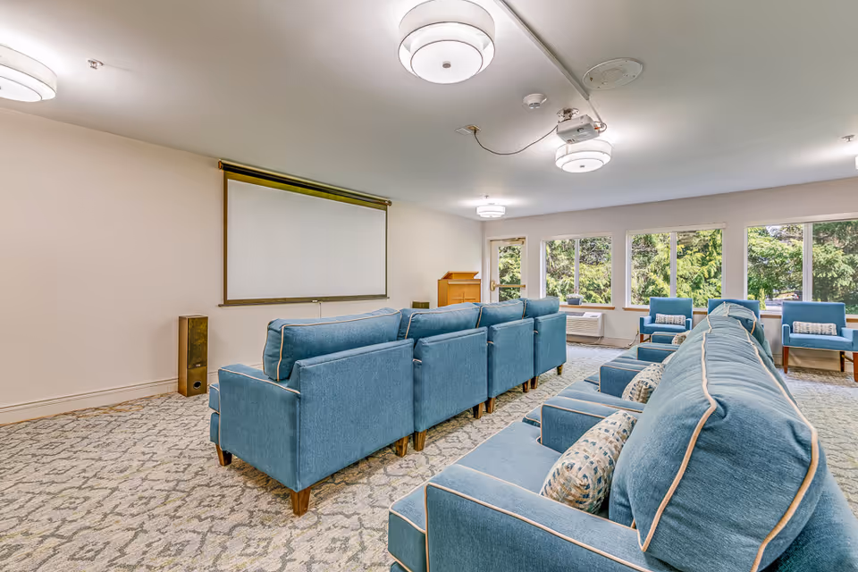 A bright senior living activity room with rows of blue upholstered chairs facing a projection screen and large windows.