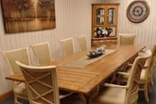 A dining room with a long wooden table surrounded by ten beige cushioned chairs. On the table is a decorative centerpiece. The walls have light-colored vertical stripes, a large clock, a wooden cabinet with glass doors, and a framed artwork.