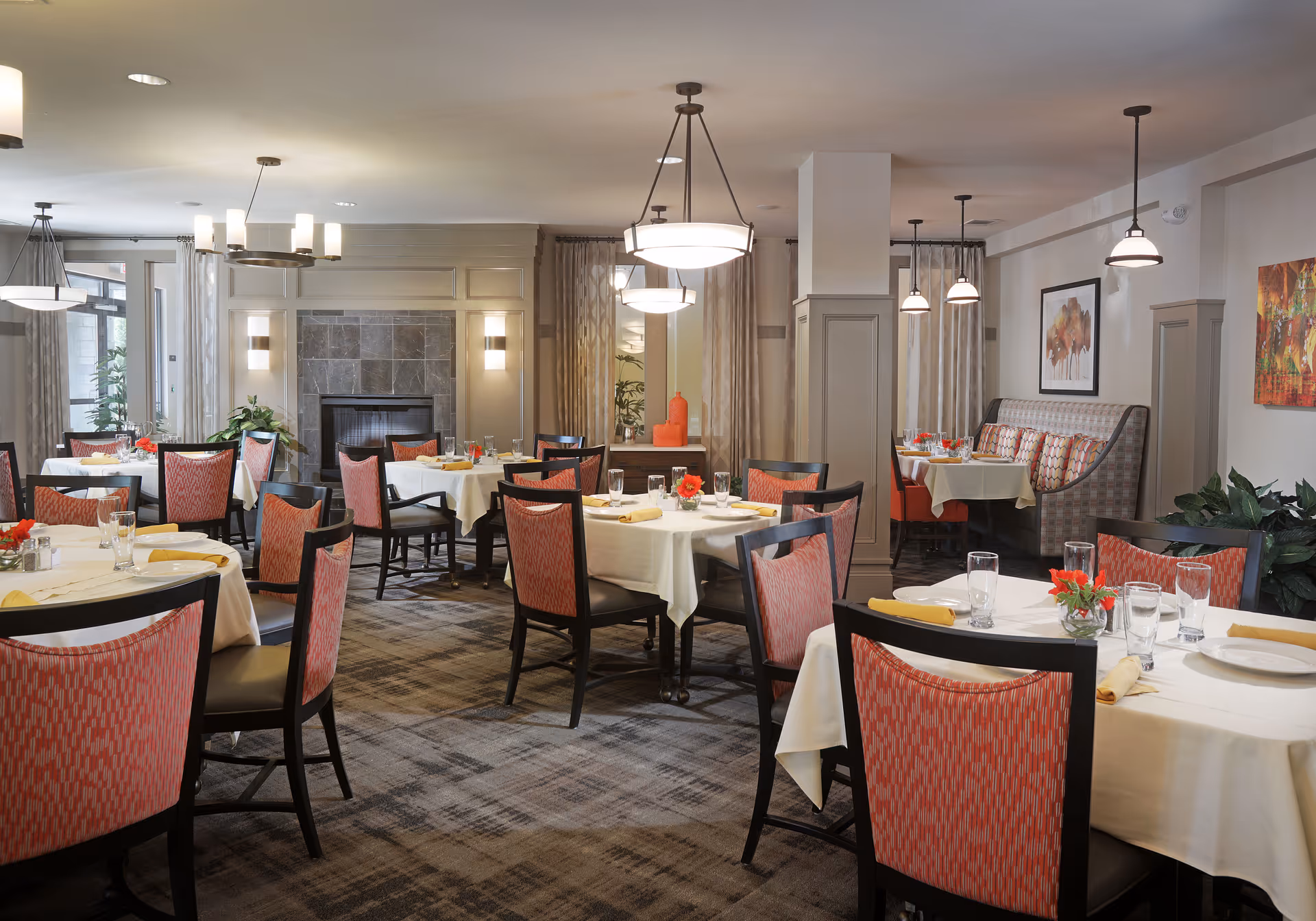 Bright, elegant dining room with multiple round tables set with white tablecloths and red-patterned chairs, pendant lights, and a central fireplace.