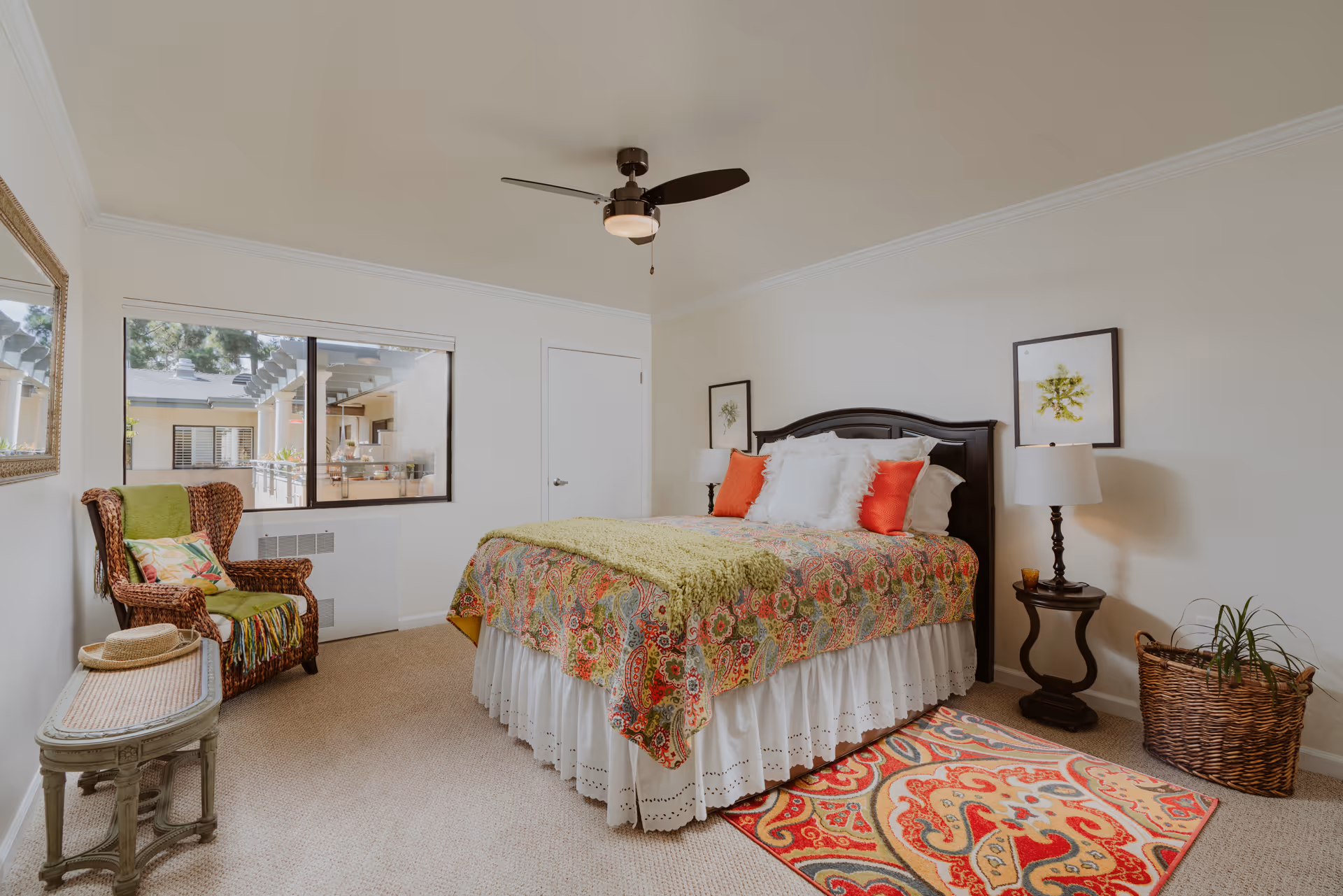 A bright and cozy bedroom with a large bed featuring a colorful floral bedspread, white and red pillows, and a green throw blanket. There is a wicker armchair with a green cushion and a colorful pillow, a small wooden table with a hat on it, a ceiling fan, two framed botanical prints on the wall, a bedside table with a lamp, a woven basket with a plant, and a vibrant patterned rug on the carpeted floor. A window shows an outdoor view of the building.