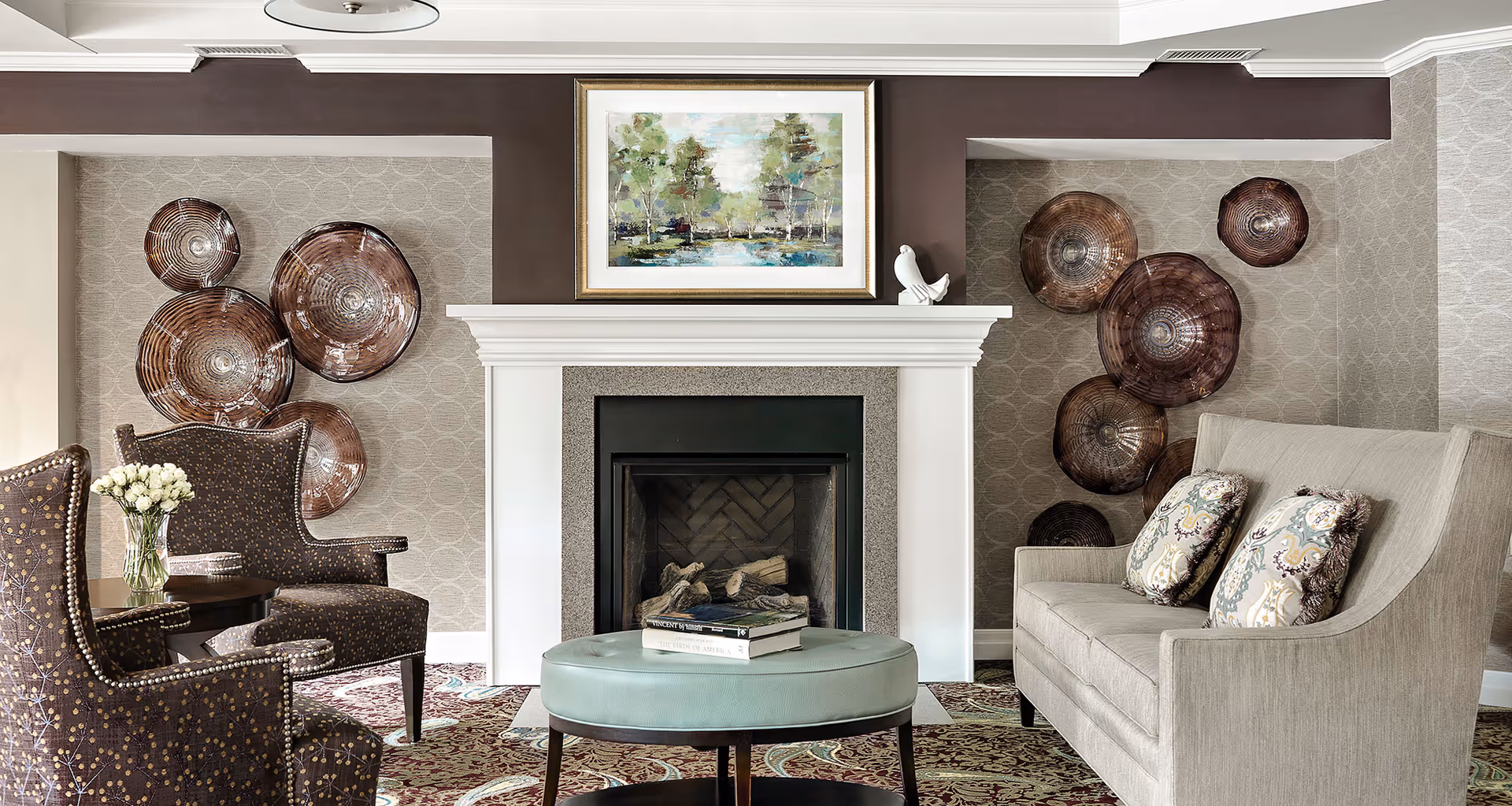 A cozy living room area featuring a white fireplace with a framed landscape painting above it. On either side of the fireplace are decorative brown circular wall hangings. The room includes two patterned armchairs with a small round table holding a vase of white flowers, a light gray sofa with two patterned pillows, and a round teal ottoman with books on top. The walls have a subtle circular patterned wallpaper and the floor is covered with a patterned carpet.