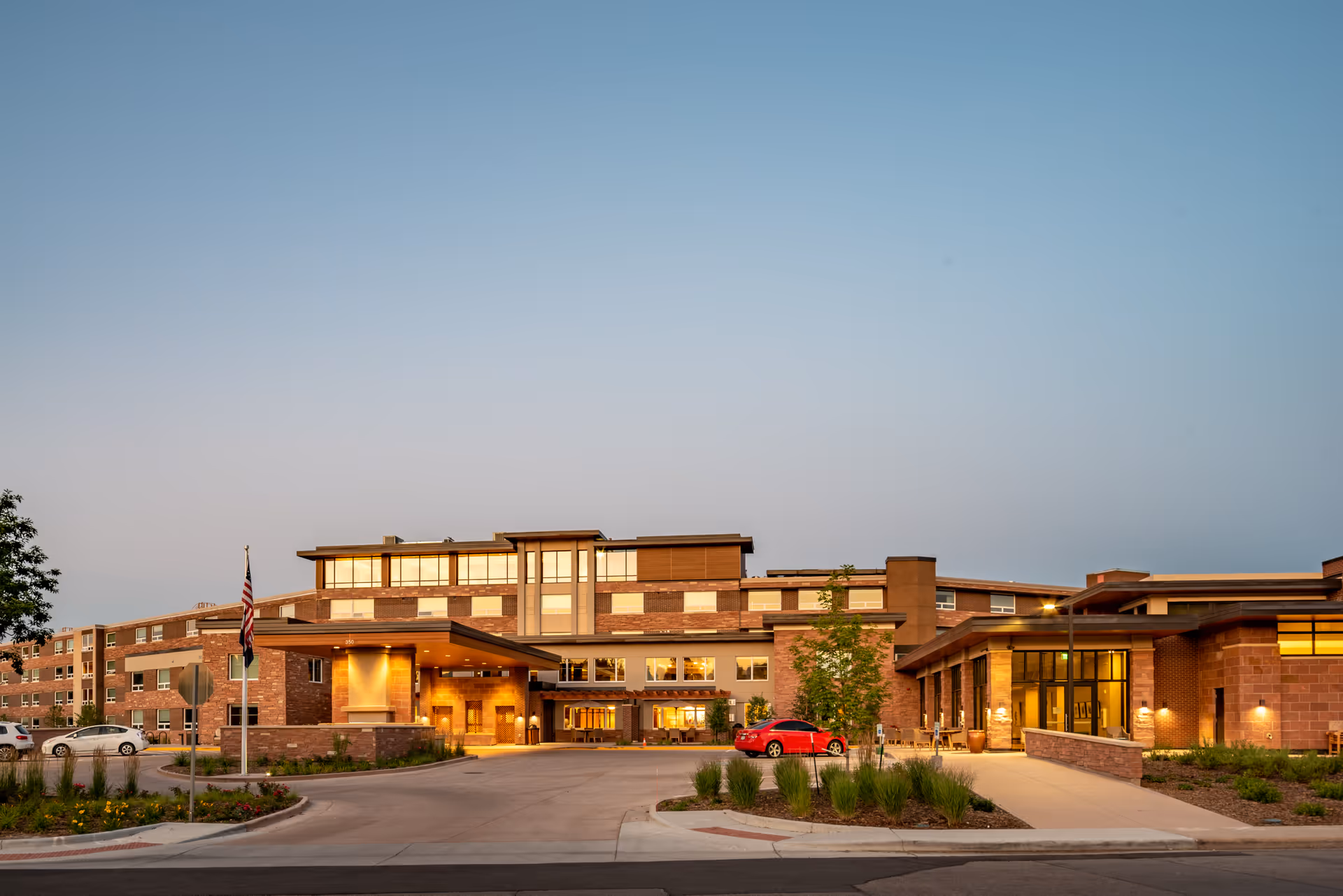 Exterior view of Frasier Retirement Community building at dusk with warm lights glowing from windows, a driveway with a red car parked, landscaped greenery, and an American flag near the entrance.