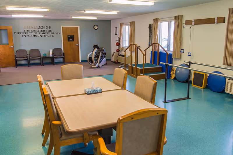 A rehabilitation room with a table and six chairs in the foreground. In the background, there are exercise equipment, parallel bars, therapy steps, and various therapy balls. The walls have windows with beige curtains and a motivational quote that reads 'CHALLENGE THE GREATER THE DIFFICULTY, THE MORE GLORY IN SURMOUNTING IT.'