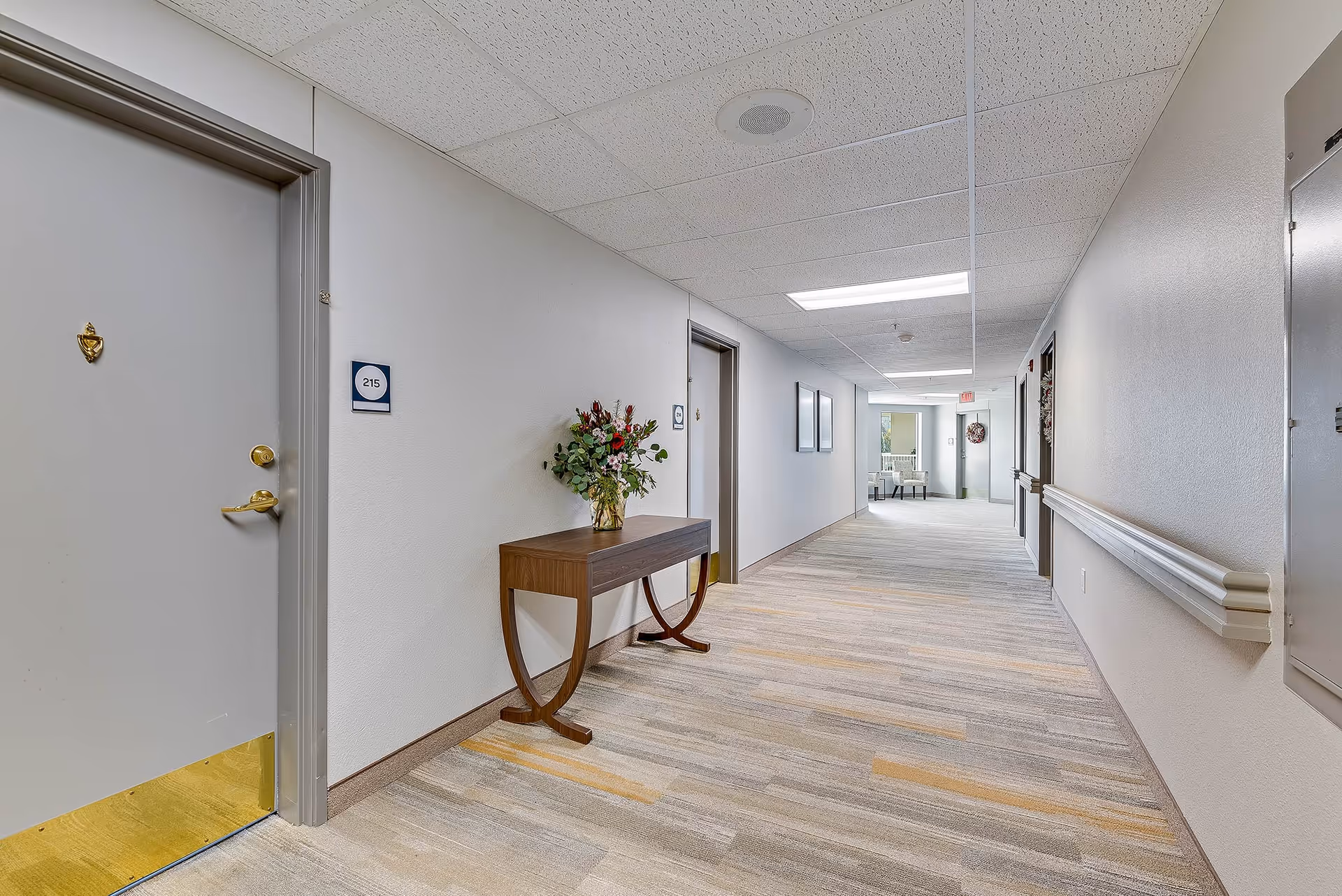 Bright, carpeted interior hallway of a senior living facility with resident doors, a console table holding flowers, handrails, and seating visible at the far end.