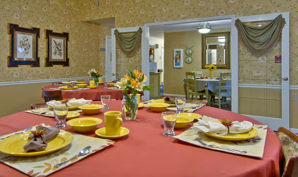 Dining room with tables covered in red tablecloths set with yellow plates, bowls, cups, and glasses. Each place setting has a napkin with a decorative flower. There are floral arrangements in glass vases on the tables. The walls have floral wallpaper and framed bird artwork. A doorway with glass double doors leads to another dining area with a table, chairs, and a mirror on the wall.
