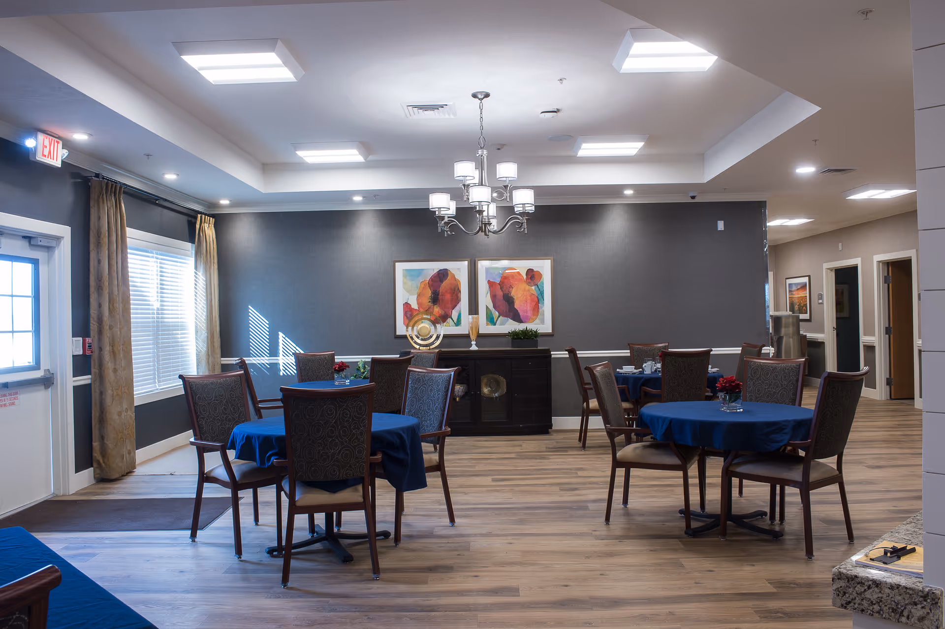 A dining room in a senior living facility with round tables covered in blue tablecloths, each surrounded by four upholstered chairs. The room has wood flooring, gray walls with white trim, two colorful abstract paintings on the back wall, and a chandelier hanging from the ceiling. There is a window with curtains on the left side and a door with an exit sign above it. The room is well-lit with ceiling lights.