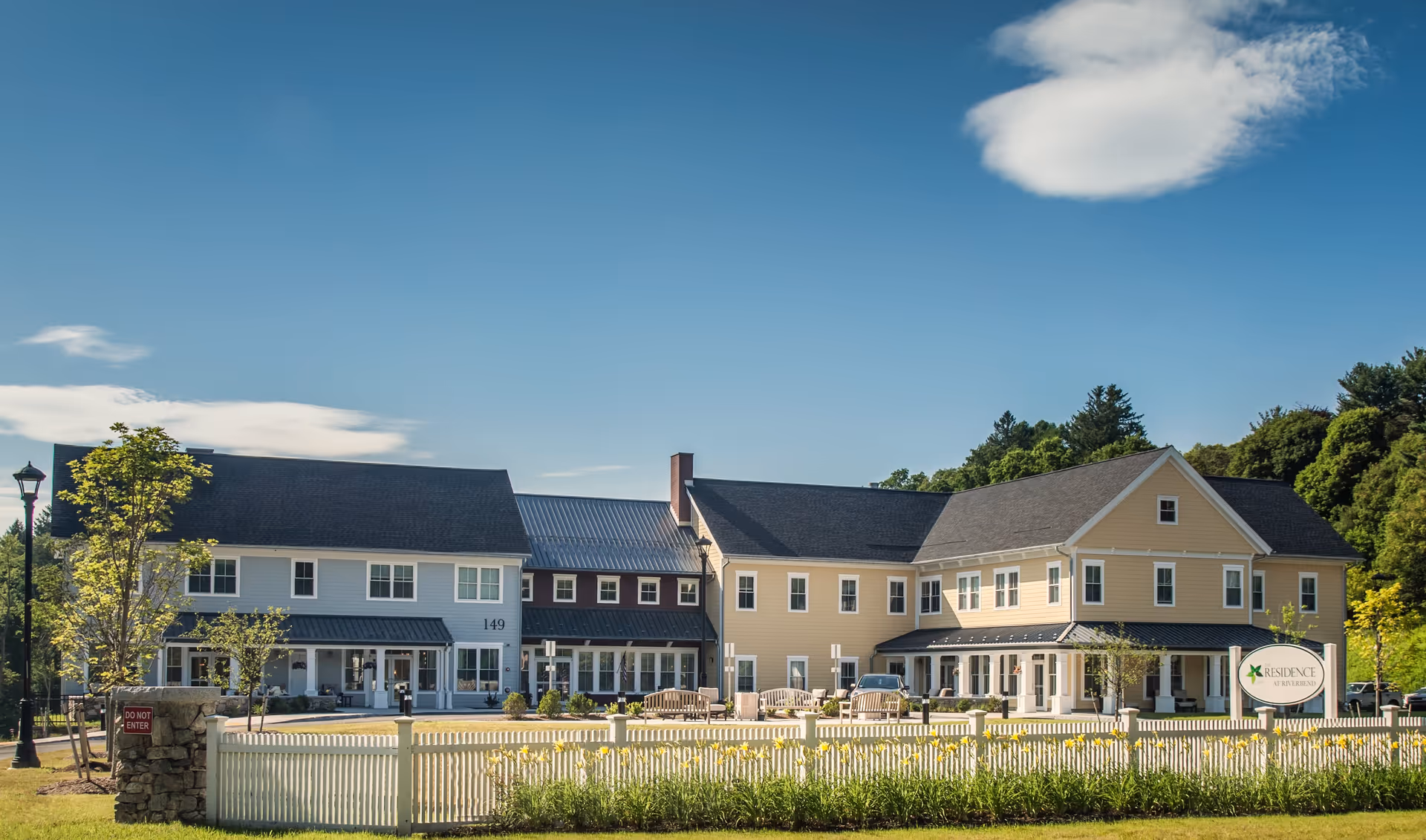 Exterior view of The Residence at Riverbend, a large two-story building with light yellow and white siding, black roofs, and multiple windows. The building is surrounded by a white picket fence, green grass, and yellow flowers. There are benches and a few small trees in front of the building under a clear blue sky with some clouds.