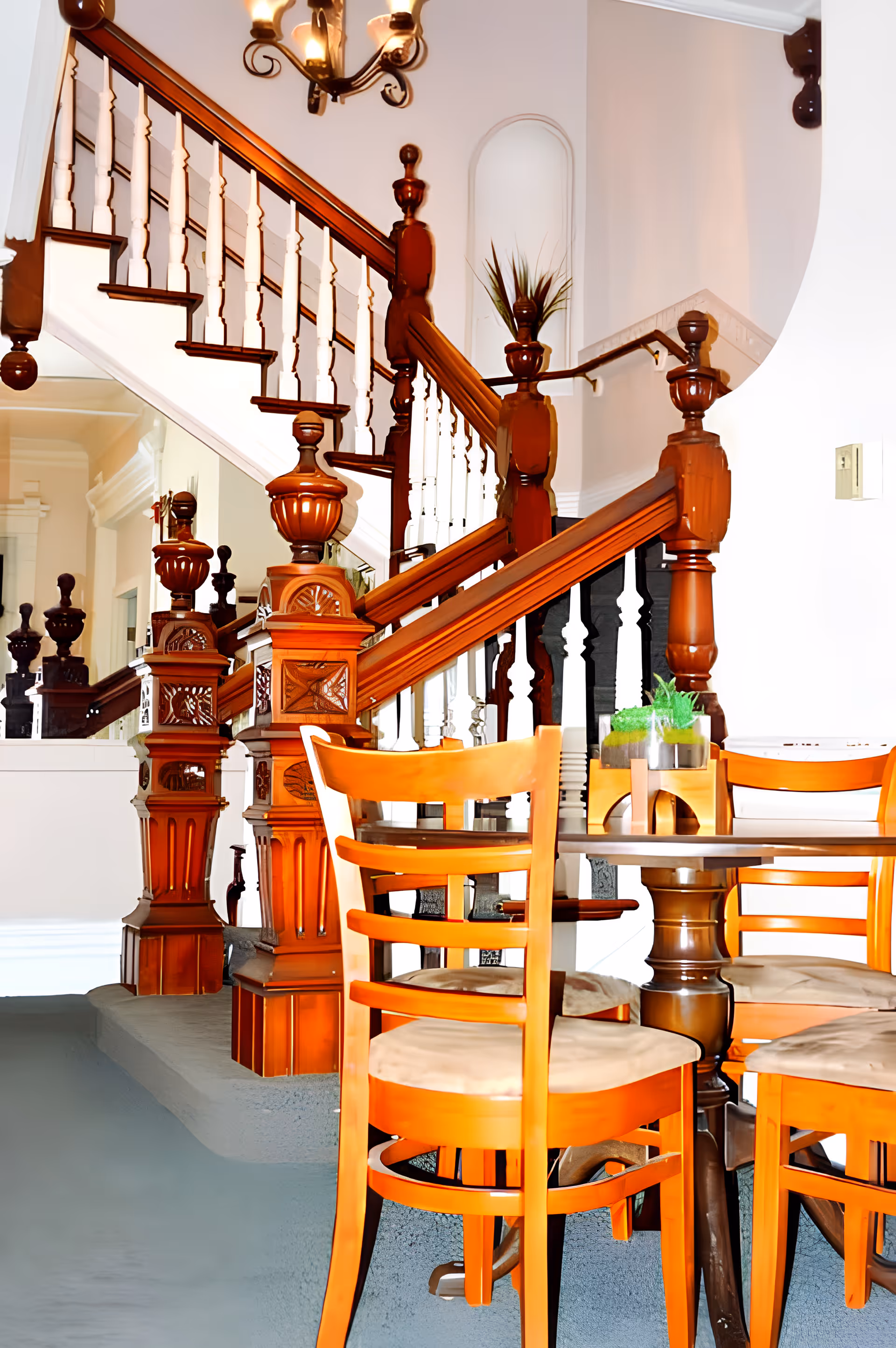 Interior view of a senior living facility showing a wooden staircase with ornate banisters and a small round table with four wooden chairs with cushioned seats. A small plant is placed on the table. The walls are light-colored, and there is a chandelier hanging above the staircase.
