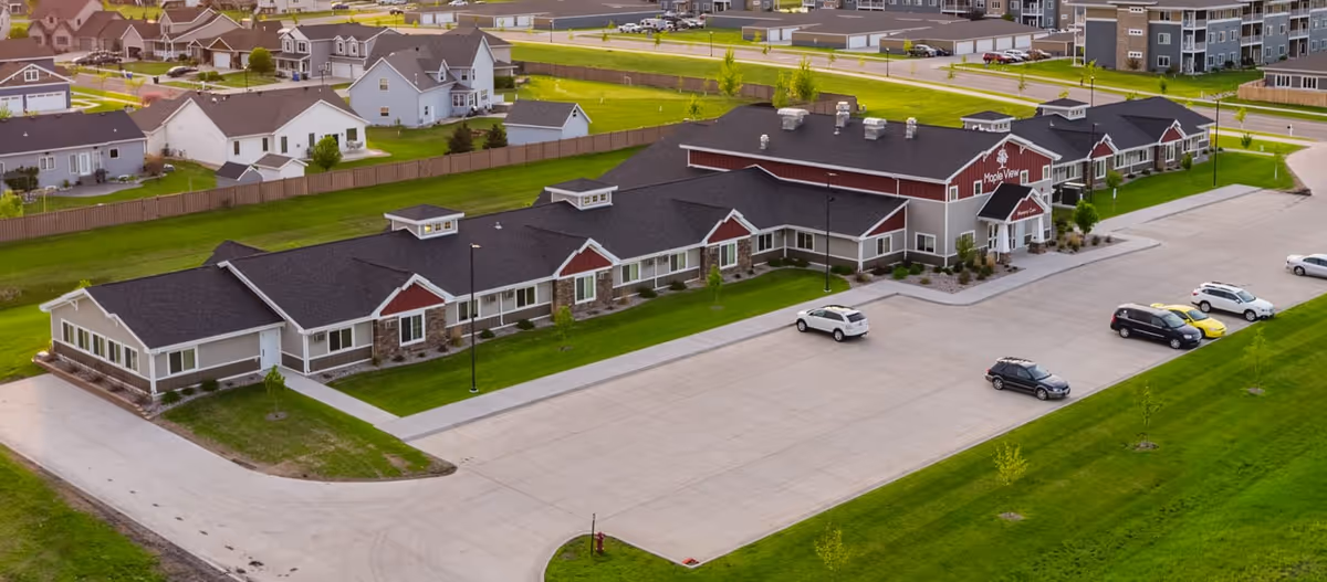 Aerial view of Fargo Maple View senior living facility showing a large single-story building with a black roof, beige and red exterior walls, and a spacious parking lot with several cars parked. Surrounding the facility are green lawns and residential houses.