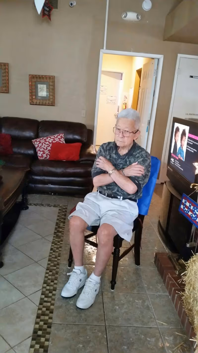 An elderly man with white hair and glasses sitting on a wooden chair in a living room. He is wearing a dark patterned shirt, light-colored shorts, white socks, and white sneakers. Behind him is a dark brown leather couch with red and patterned cushions. The room has tiled flooring with a decorative border, beige walls with framed artwork, and a TV on a stand to the right displaying a program with two people on screen.
