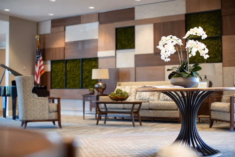 A well-lit living room area with a round table in the foreground holding a pot of white orchids. In the background, there is a beige sofa, two armchairs, a coffee table with decorative items, a table lamp, and an American flag. The wall behind features a modern design with wood panels and green moss squares.