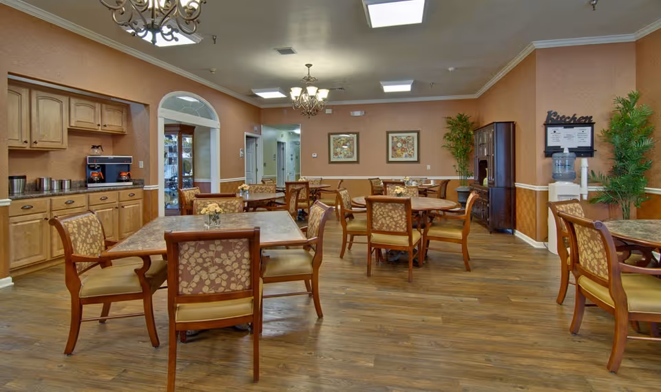 A dining room in a senior living facility with several wooden tables and chairs arranged neatly. The room has warm-toned walls, wooden flooring, and decorative plants. There are chandeliers hanging from the ceiling and framed floral artwork on the walls. A water dispenser and a cabinet are visible in the background.
