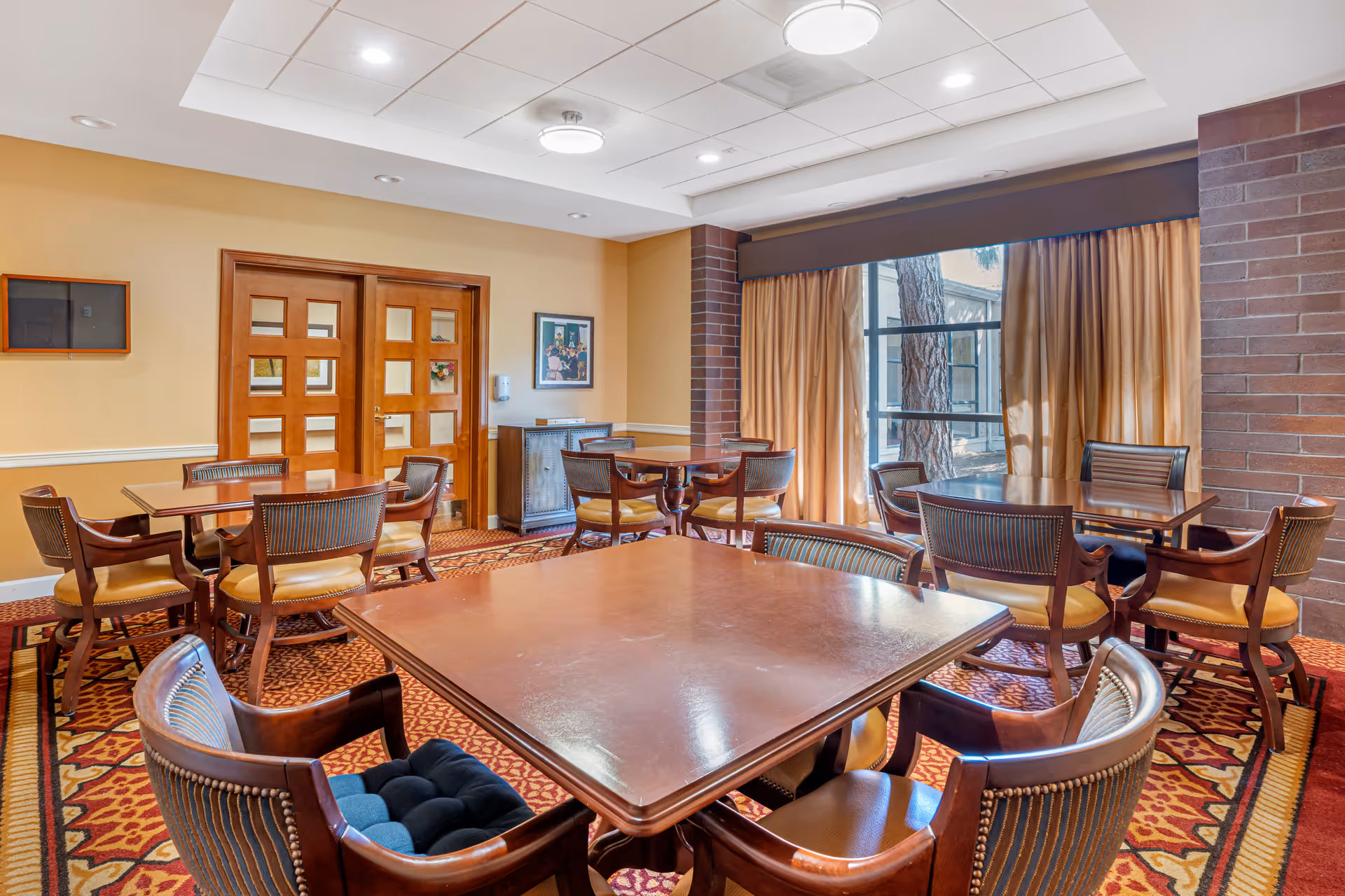 Interior view of a senior living facility dining room with several wooden tables and chairs arranged on a patterned carpet. The room has beige walls, a large window with beige curtains, and double wooden doors. A framed picture and a small cabinet are visible against the wall.