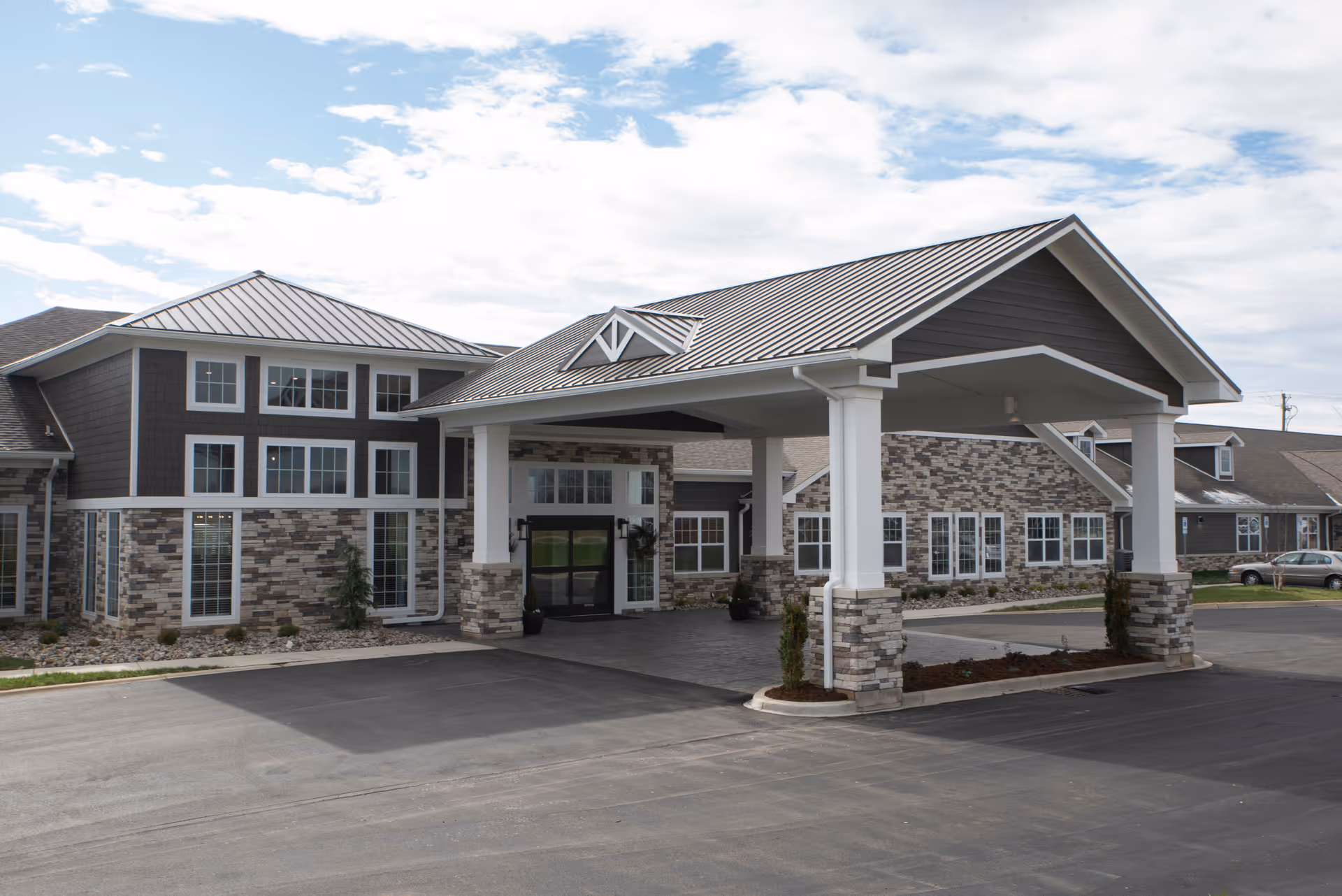 Front entrance of a senior living building with a covered drop-off canopy, stone facade, and multiple windows.