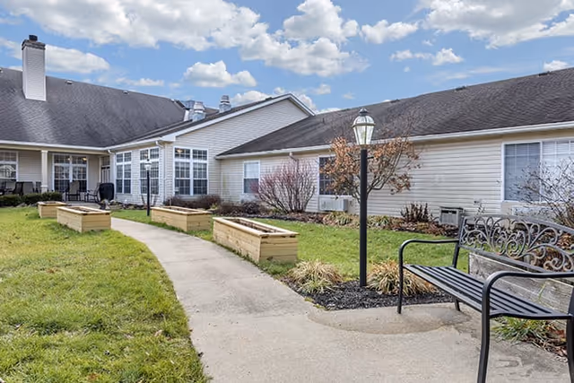 Outdoor courtyard area of a senior living facility with a concrete walkway, raised garden beds, a black metal bench, a lamp post, and a building with white siding and multiple windows under a partly cloudy sky.