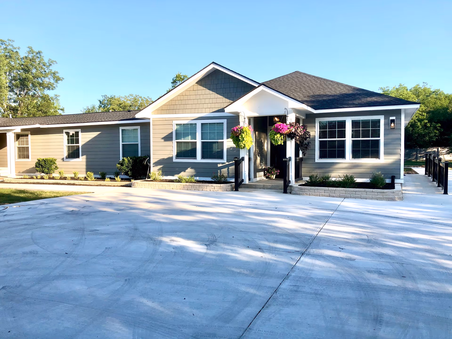 Exterior view of a single-story senior care facility building with gray siding, white trim, and a dark shingled roof. The entrance features a small porch with hanging flower baskets and black railings. There are several windows with white frames and a concrete driveway in front. Trees and greenery surround the building under a clear blue sky.