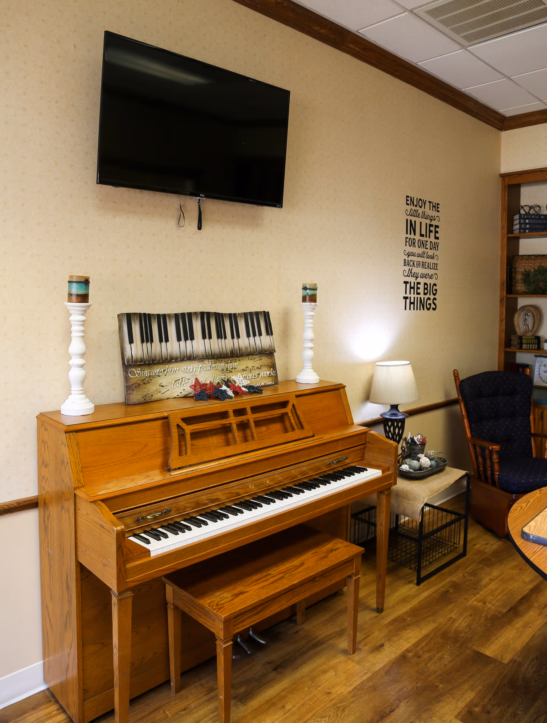 A cozy room featuring a wooden upright piano with a matching bench, two white candle holders with candles on top of the piano, a decorative piano-themed sign, a wall-mounted flat-screen TV above the piano, a small side table with a lamp and decorative items, a cushioned rocking chair, and a bookshelf in the corner. The wall has a motivational quote decal.