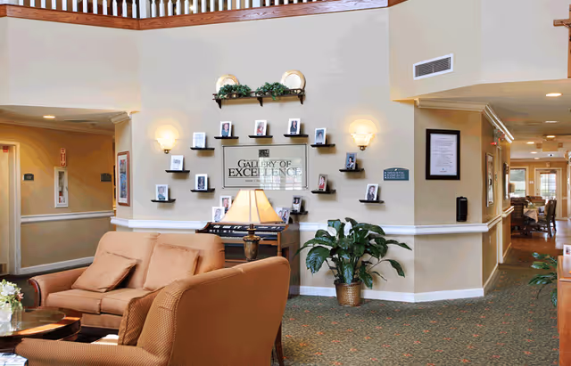 A cozy living room area in Wellington Manor Knoxville featuring a brown sofa and armchair, a small table with a lamp, a plant in a pot, and a wall display titled 'Gallery of Excellence' with framed photos. The space has beige walls, carpeted flooring, and a railing balcony above.