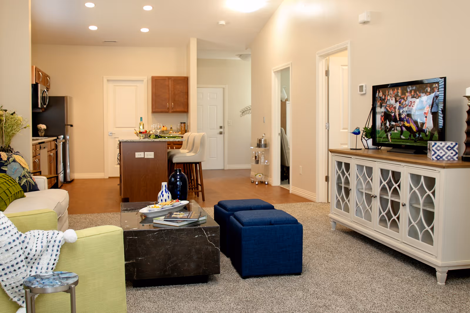 Open-plan living room and kitchen with a sofa, coffee table, blue ottomans, TV on a console, and a kitchen island with barstools.