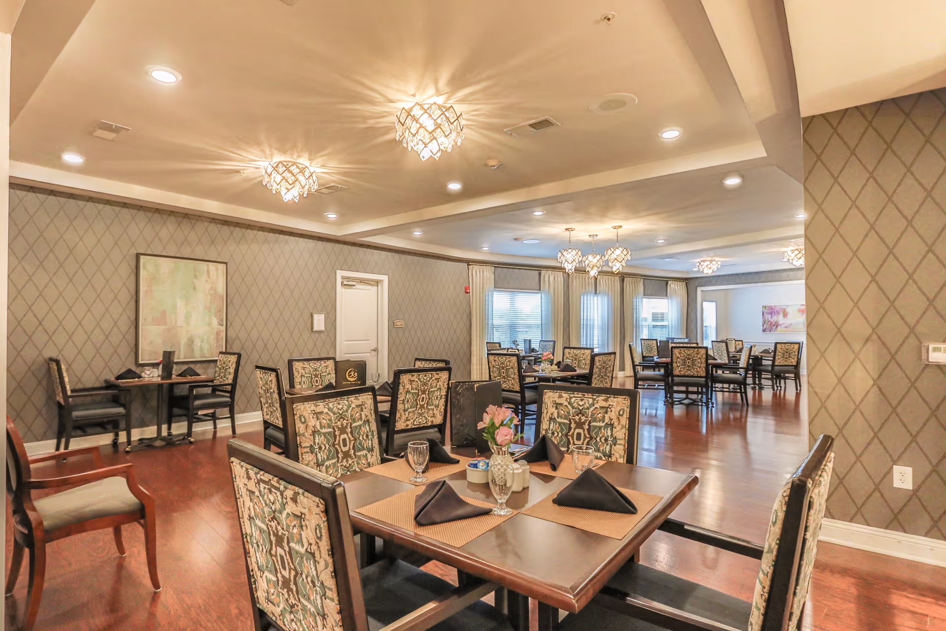 Dining room with multiple set tables, patterned chairs, chandeliers, and hardwood floors in a senior living facility.