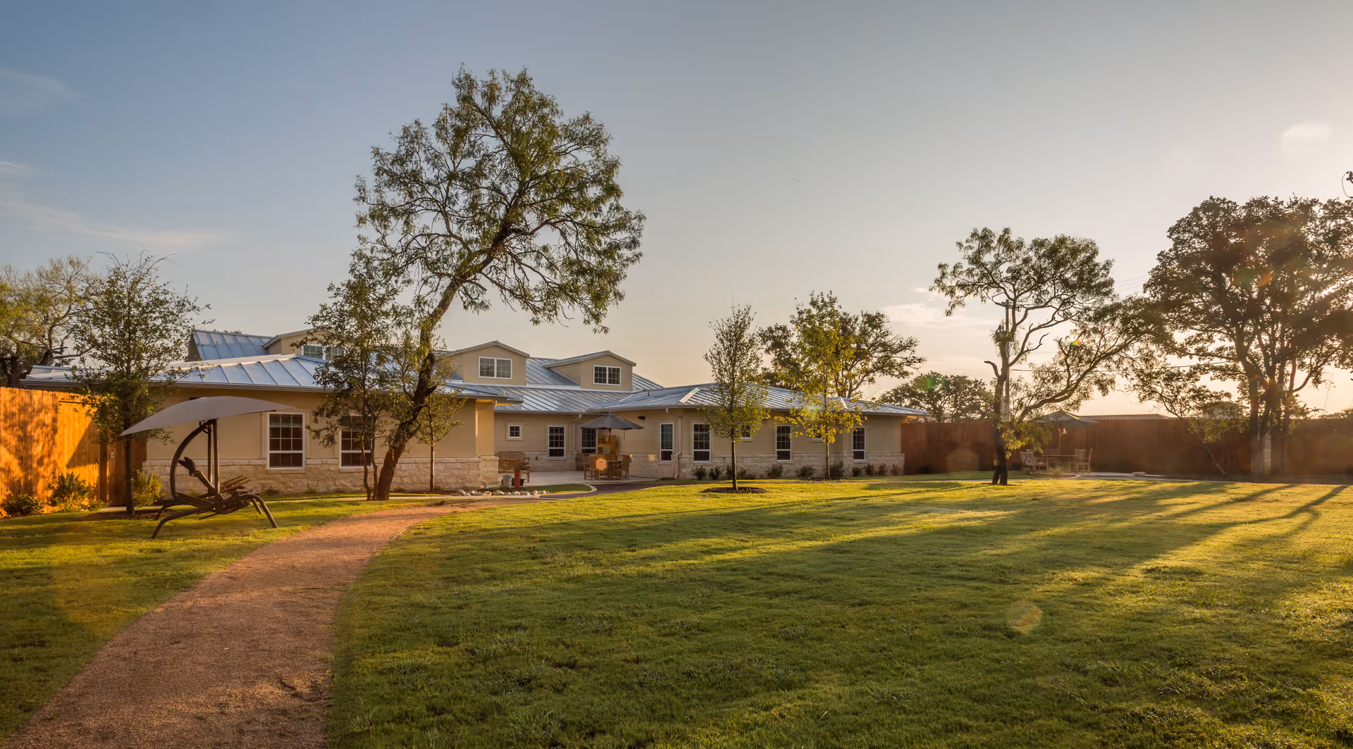 A single-story building with a metal roof surrounded by a large grassy yard with several trees and a curved walking path. There is a shaded outdoor lounge chair on the left side and a wooden fence enclosing the area. The scene is lit by warm sunlight, suggesting either early morning or late afternoon.