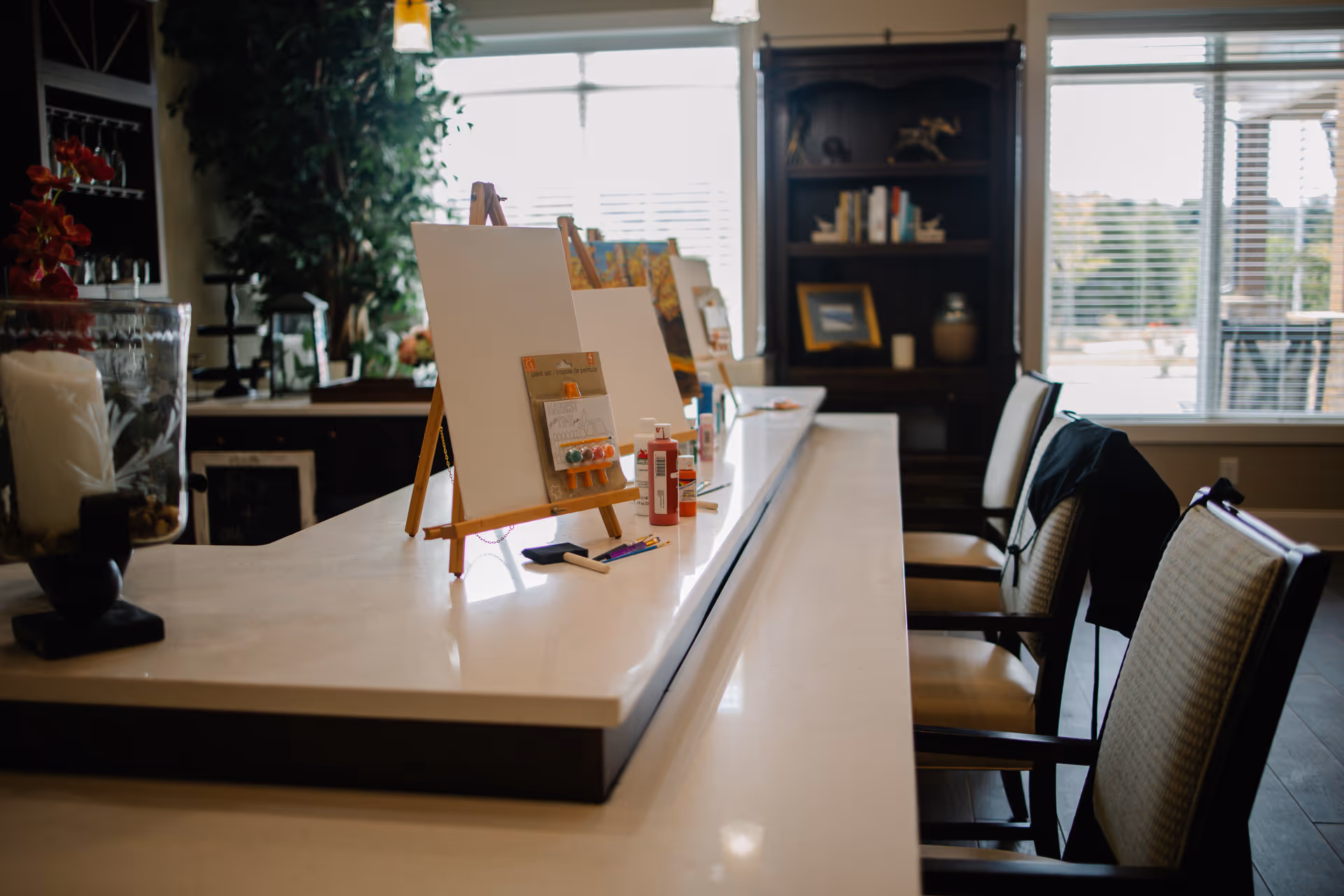 A bright room with a long white countertop featuring several small easels holding blank canvases and painting supplies like brushes and paint bottles. There are cushioned chairs lined up along the counter, and large windows in the background letting in natural light. Shelves with books and decorative items are visible in the background.