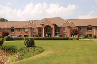 Front exterior view of a two-story brick building with a central entrance featuring columns, surrounded by well-maintained green lawns and small trees under a partly cloudy sky.