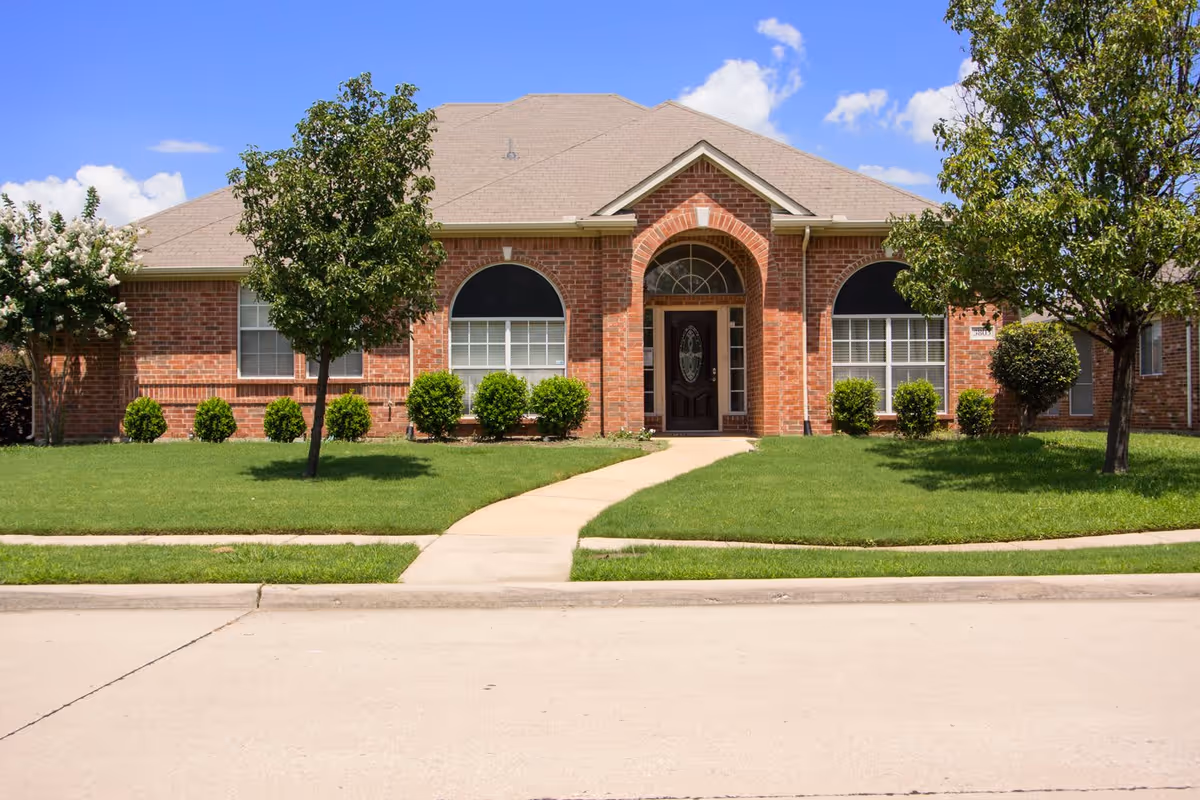 Front exterior view of a single-story brick building with a peaked roof, arched doorway with a decorative glass front door, two large windows with black awnings, green lawn, and trees on either side of a concrete walkway leading to the entrance under a blue sky with some clouds.