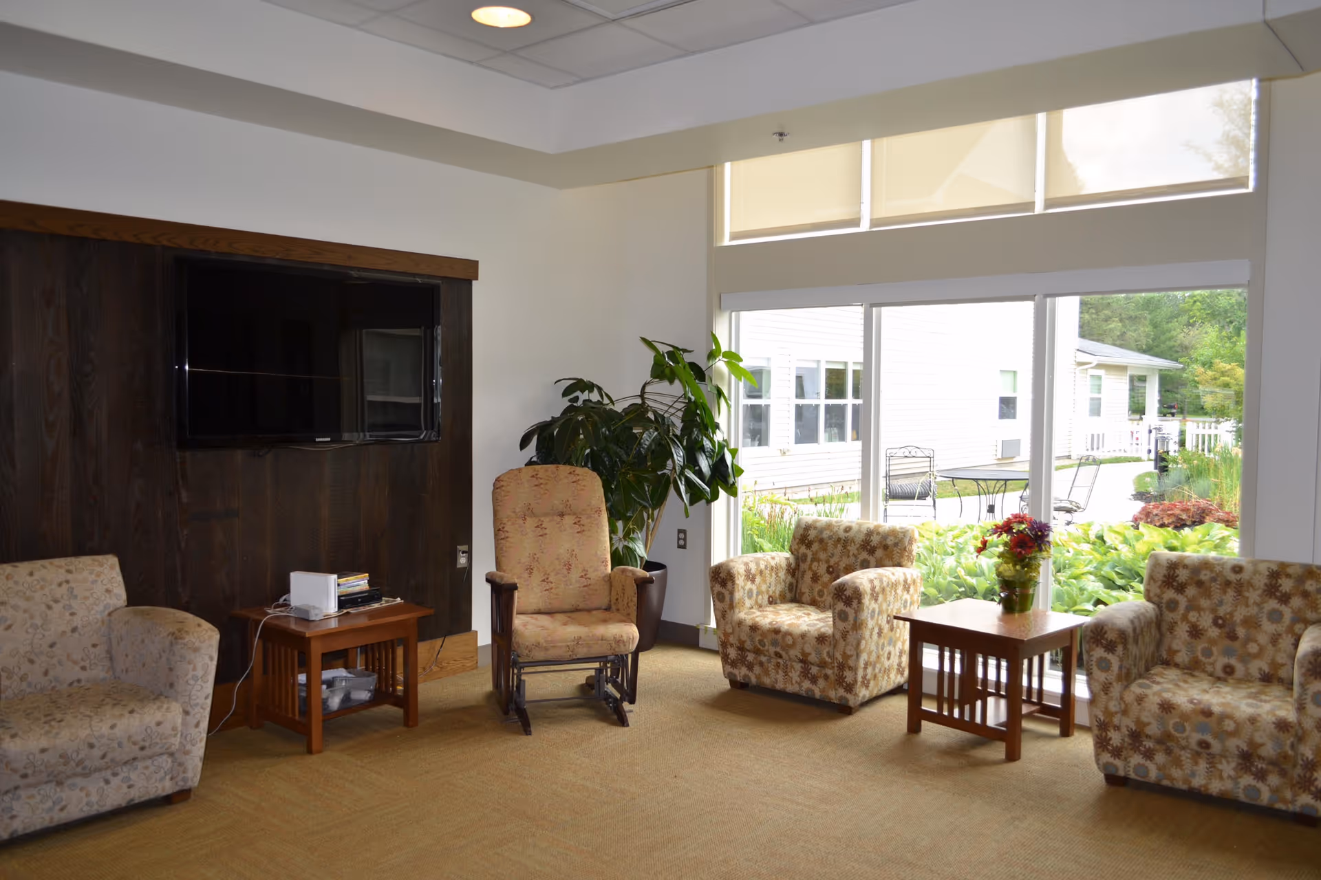A cozy living room area with four upholstered armchairs arranged around two wooden side tables. A large flat-screen TV is mounted on a dark wood-paneled wall. There is a large window letting in natural light and showing greenery and outdoor seating outside. A potted plant is placed near the window.