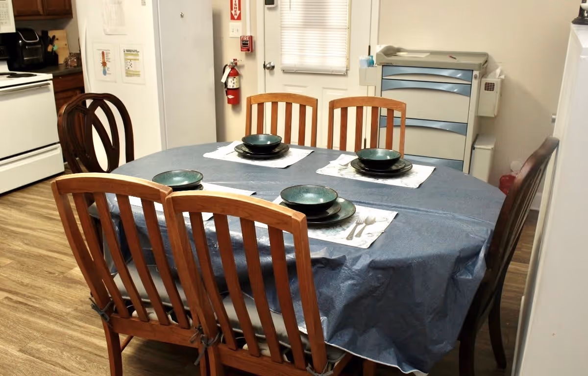 A dining area with a rectangular table covered by a blue tablecloth. The table is set with four place settings, each including a bowl, a plate, and silverware on a placemat. Surrounding the table are six wooden chairs with vertical slats. In the background, there is a white door, a fire extinguisher mounted on the wall, a white stove, wooden cabinets, and a multi-drawer storage unit.