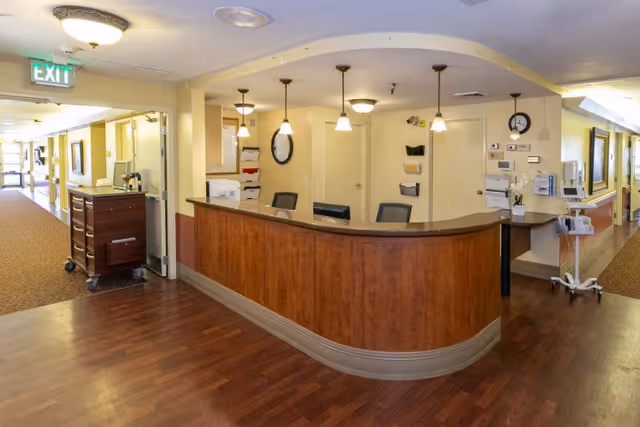 Reception area inside Holladay Healthcare Center featuring a curved wooden front desk with three chairs behind it, overhead pendant lights, and a hallway extending to the left and right. The floor is wood laminate, and there are various office supplies and wall-mounted organizers visible behind the desk.