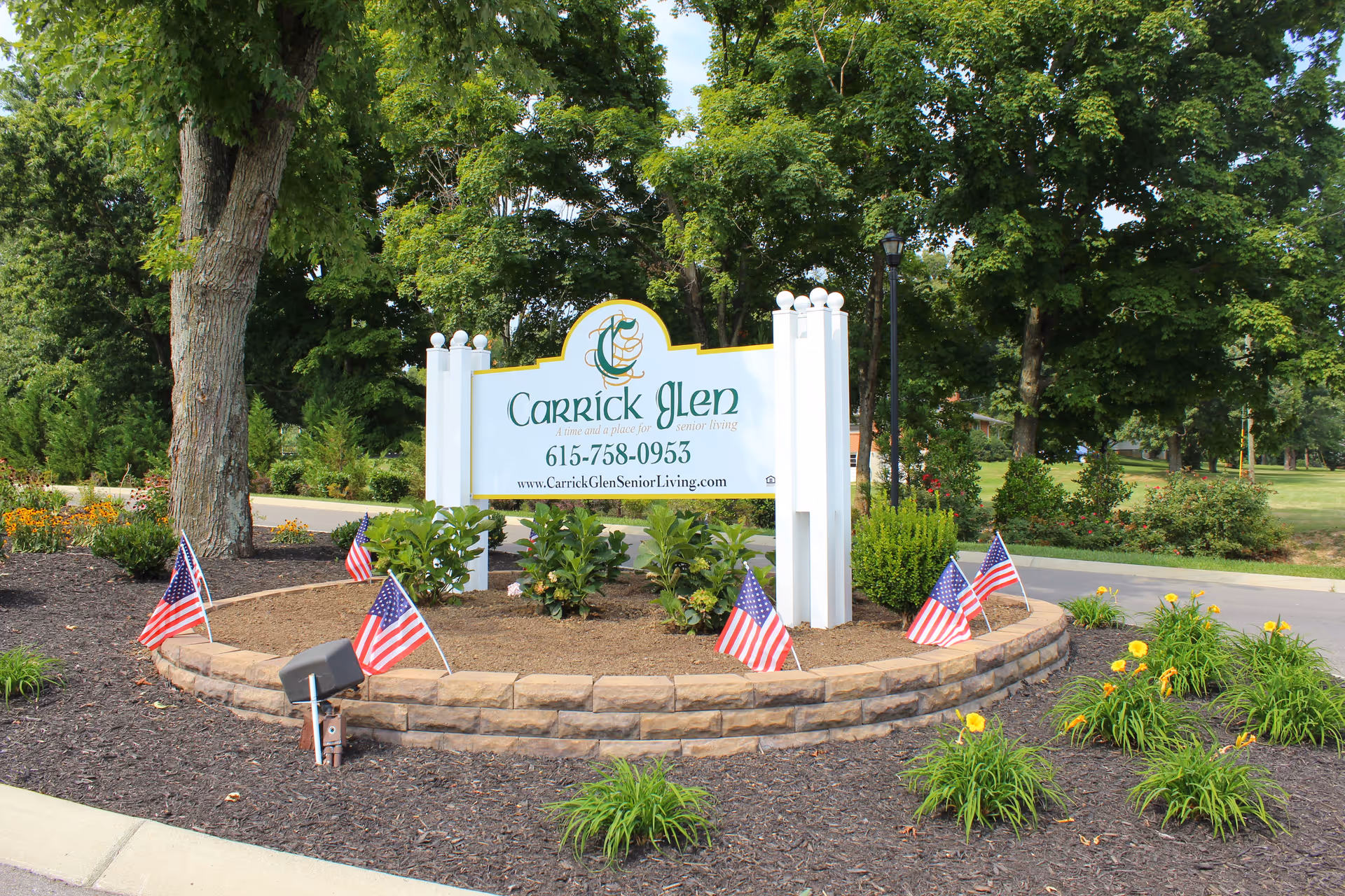 Outdoor landscaped area with a white sign for Carrick Glen Senior Living surrounded by small American flags, plants, and trees in the background.