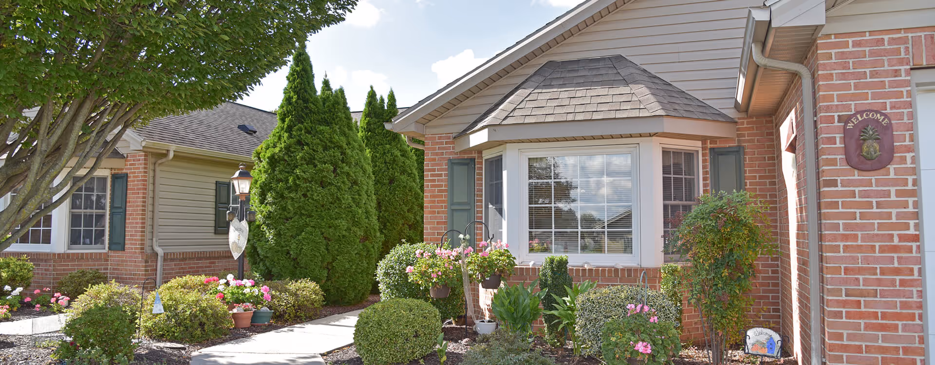 Exterior view of a brick and siding building with a bay window, green shutters, and a landscaped garden featuring bushes, flowers, and a small tree. A concrete walkway leads through the garden, and a decorative welcome sign is mounted on the brick wall near the entrance.