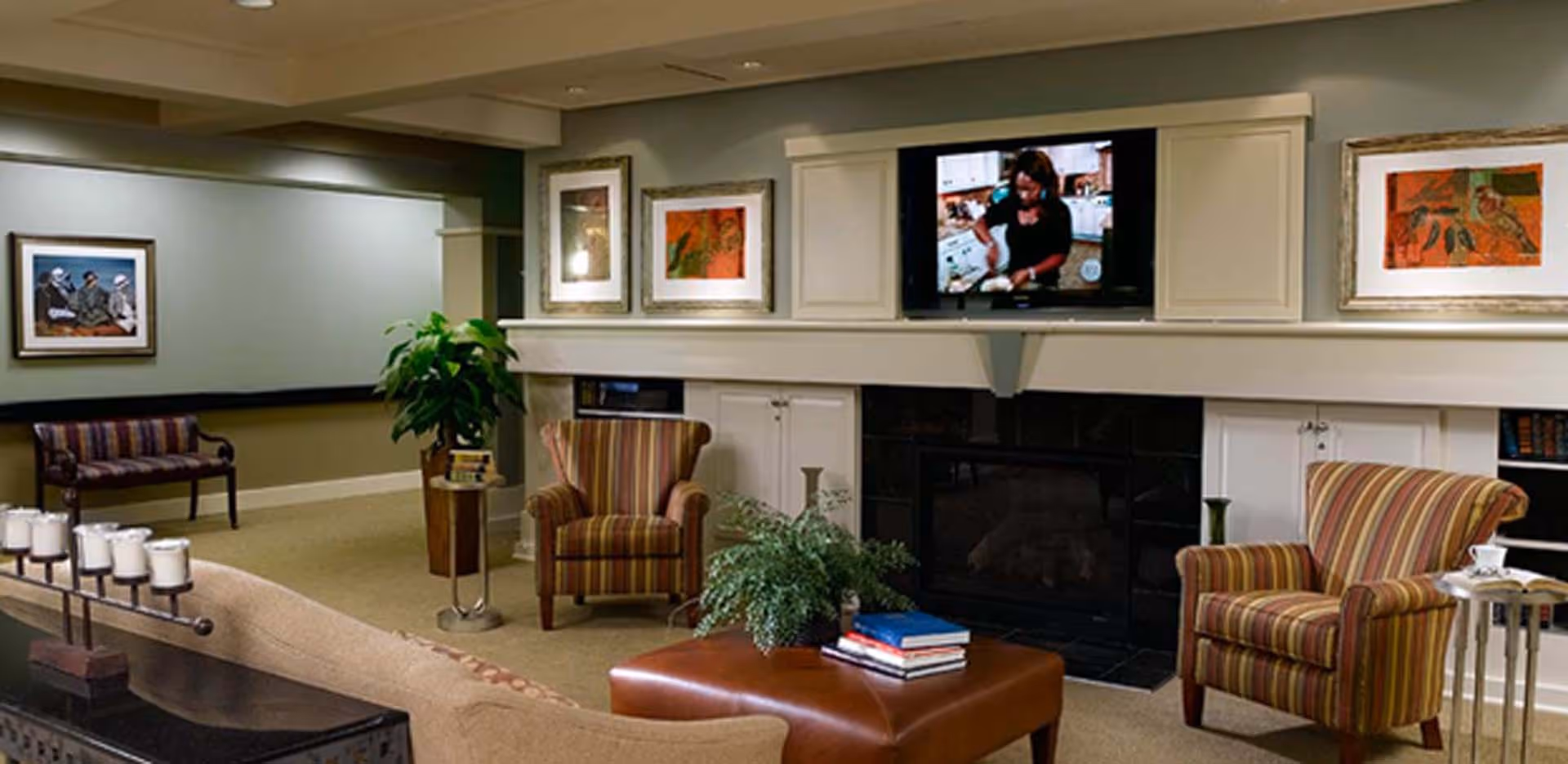 Comfortable communal living room with striped armchairs, a sofa and ottoman, a TV above a fireplace, framed artwork, and plants.