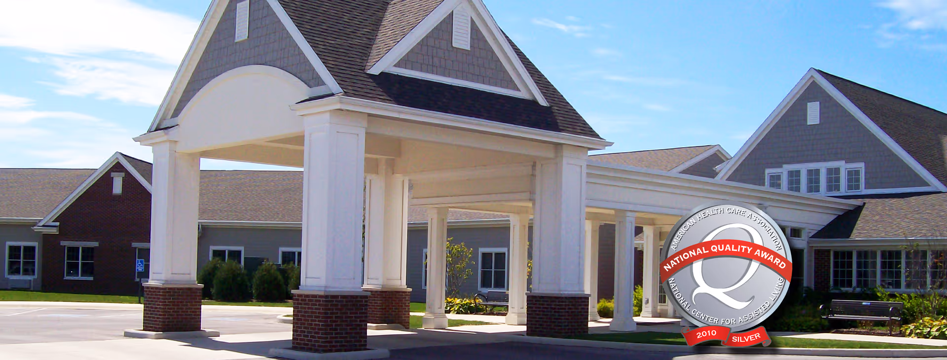 Exterior view of Briarwood Village senior living facility showing a covered entrance with white columns and brick bases, connected to the main building with gray siding and multiple windows under a clear blue sky.