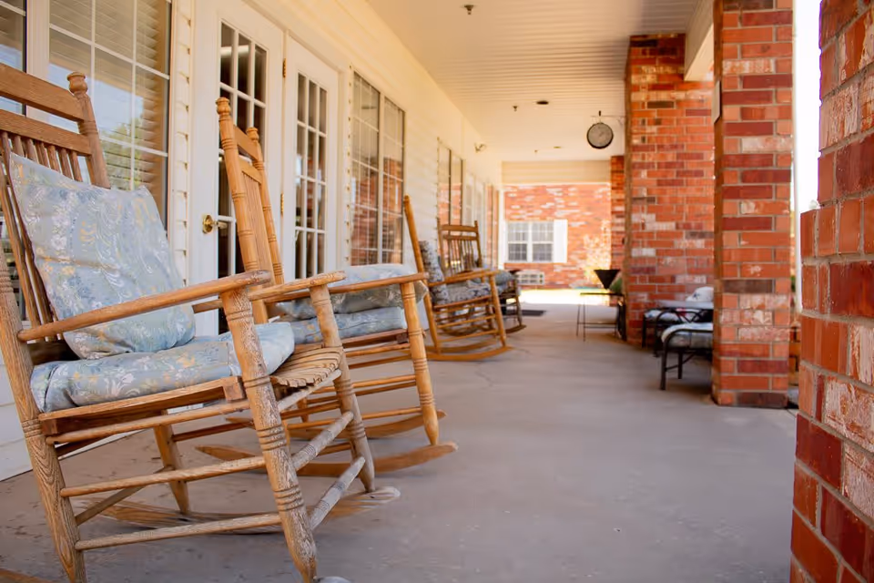 A covered outdoor porch area with wooden rocking chairs that have floral cushions, brick pillars, and glass doors leading inside. There is a clock hanging from the ceiling and additional seating further down the porch.