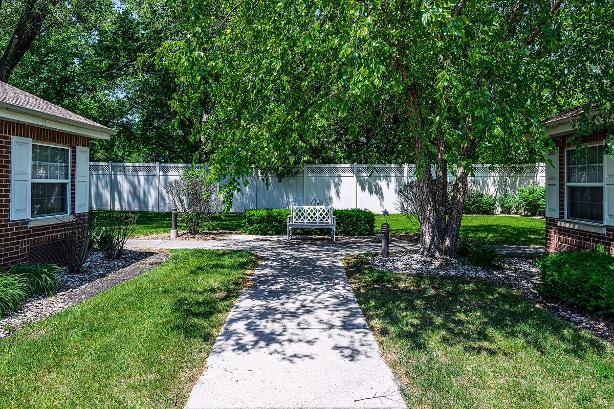 A peaceful outdoor garden area with a concrete walkway leading to a white bench under a large leafy tree. The area is surrounded by green grass, bushes, and two brick buildings with white window shutters on either side. A white privacy fence encloses the garden space.