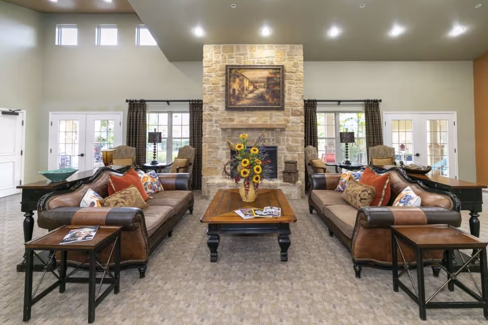 A spacious and well-lit living room with two brown leather sofas facing each other, each adorned with colorful throw pillows. A wooden coffee table with a vase of sunflowers and magazines sits between the sofas. Behind the coffee table is a stone fireplace with a framed painting above it. Large windows with dark curtains and glass doors allow natural light to fill the room. Additional seating with armchairs and side tables are placed near the windows.