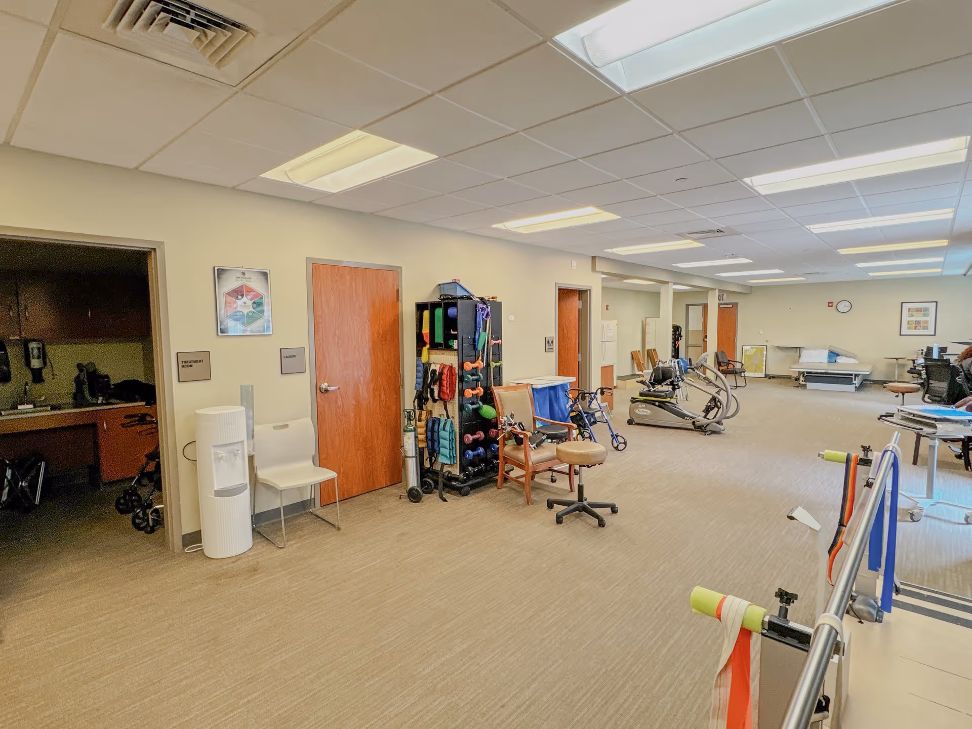 A spacious therapy and exercise room at VibraLife, featuring various exercise equipment, chairs, and a water cooler. The room is well-lit with overhead lighting and has a neutral color scheme.